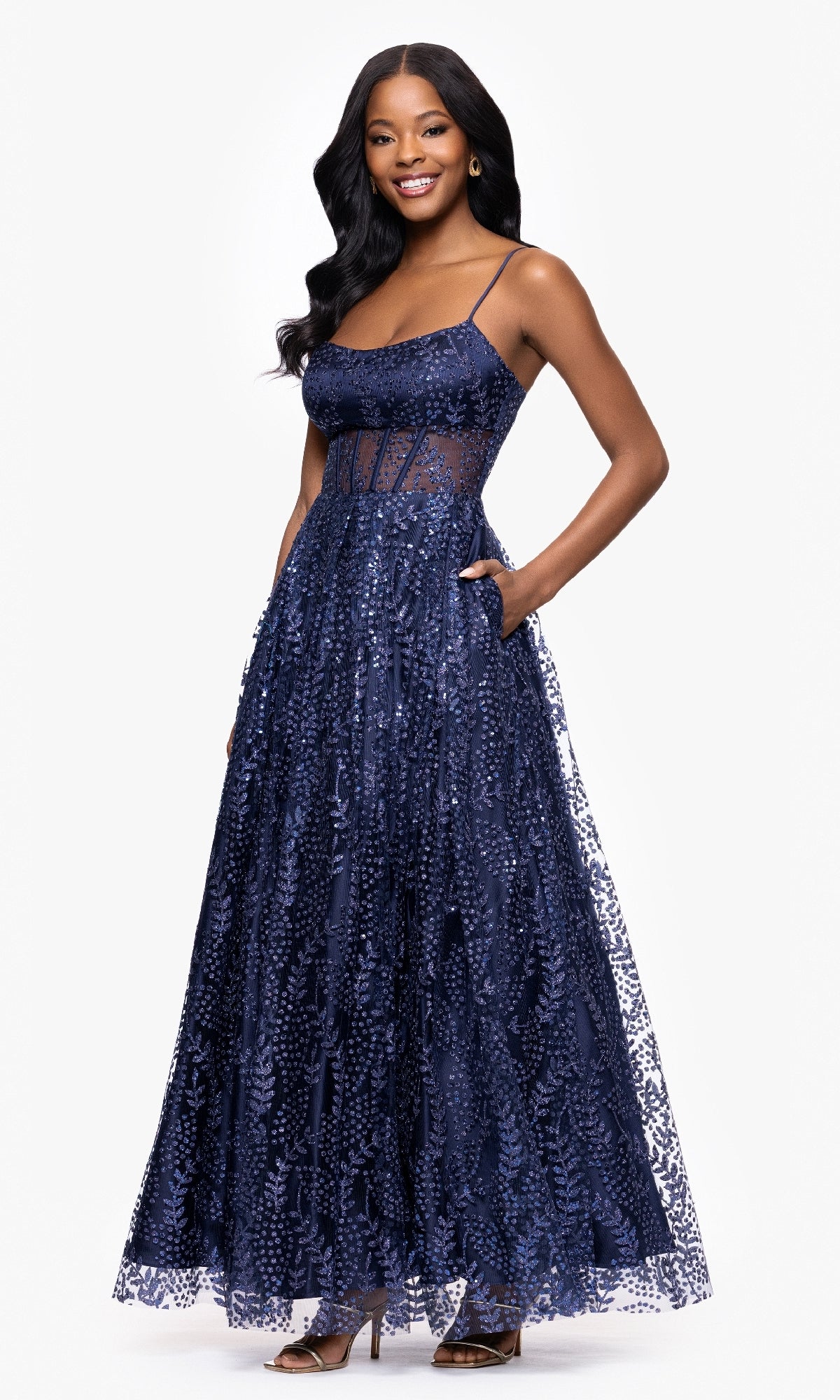 Smiling woman in a navy Glitter-Print Prom Ball Gown (Blondie Nites 5539BN) with spaghetti straps, sheer bodice detailing, and sequin embellishments, standing against a white background.